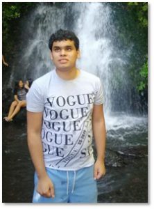 A photo of a young dark wavy hair man in a printed T-shirt standing in front of a waterfall.